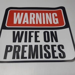 May include: A rectangular sign with a red and white "WARNING" banner at the top. Below, the sign reads "WIFE ON PREMISES" in bold black letters on a white background, framed by a black border. The sign is likely for a home or business.