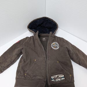 May include: Brown quilted zip-up hooded jacket with a dark blue fleece-lined hood. Features a "Rural Road Adventure Trails" patch and a circular patch with text. The jacket has two front pockets and a small red patch on the left sleeve.