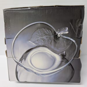 May include: A clear glass apple-shaped serving dish with a leaf detail, displayed on a box with a matching image. The dish has a stem and a smooth, curved design. The box is gray and black.