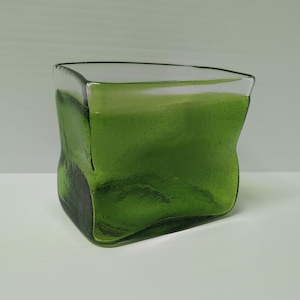 May include: A green glass candle holder with a distinctive, slightly irregular form. The glass is a deep, rich green, and the candle holder is filled with green wax. The design is modern and minimalist.
