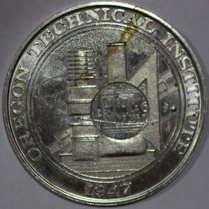May include: A silver-colored coin featuring the text "OREGON TECHNICAL INSTITUTE" around the edge. The center design includes a building, a circle with a smaller building inside, and the year "1947" at the bottom.