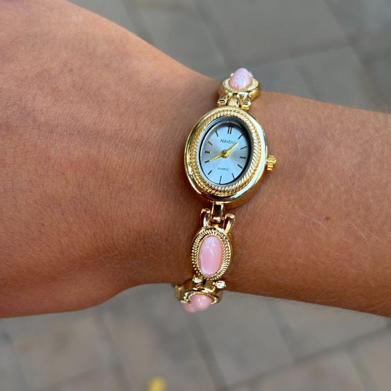 May include: Gold-toned wristwatch with an oval face and a light blue dial. The watch features a gold band with pink oval-shaped accents. The watch face has the word "NAIDU" printed on it.
