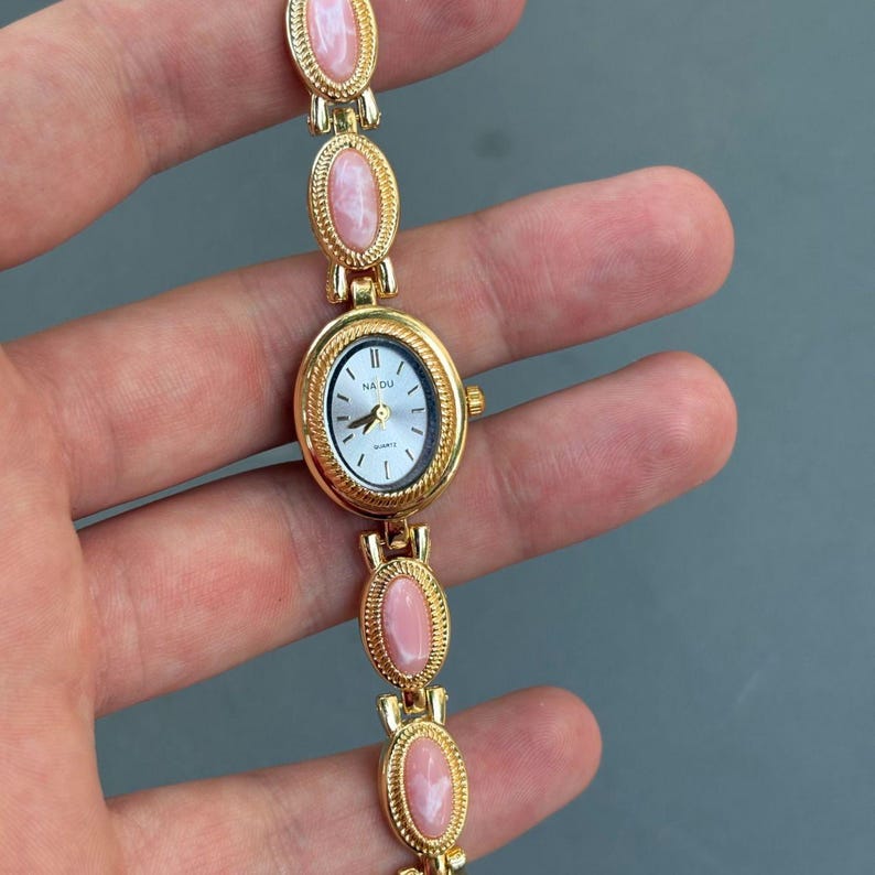May include: A gold-toned wristwatch with a white face and pink oval accents. The watch face has black hands and the text "NA DU". The watch band features a series of pink oval stones set in gold-colored metal.