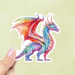 May include: A watercolor sticker of a colorful dragon. The dragon has rainbow wings and a body with shades of blue, purple, and red. The sticker is held by a hand with a light pink nail.