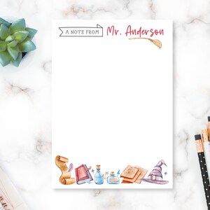 May include: White notepad with the text "A NOTE FROM Mr. Anderson" in black and red. The notepad features watercolor illustrations of a scroll, books, potion bottles, a witch hat, and a broom. Pencils and a succulent plant are visible.