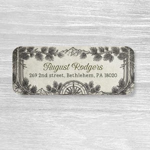 May include: A rectangular address label featuring a black ink illustration of pine trees and mountain peaks framing the text August Rodgers, 269 2nd street, Bethlehem, PA 18020, with a compass rose graphic centered at the bottom edge of the label.
