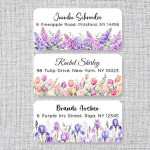 May include: Three rectangular address labels with rounded corners. The top label features purple lilacs and the name Jannika Schroeder with address 8 Pineapple Road, Pittsford, NY 14456. The middle label shows colorful tulips and the name Rachel Shirley with address 98 Tulip Drive, New York, NY 10023. The bottom label displays blue and purple irises and the name Brandi Archer with address 6 Purple Iris Street, Riga, NY 12345.