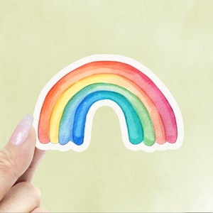 May include: A colorful rainbow sticker with a white border. The rainbow features bands of red, orange, yellow, green, blue, and purple. The sticker is held by a person's hand against a light green background.