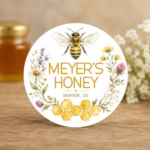 Può includere: Un'etichetta circolare bianca con un'illustrazione centrale di un'ape sopra il testo dorato MEYER'S HONEY e DENVER, CO. Il design include una ghirlanda floreale di margherite, lavanda e trifoglio, con celle di alveare gialle in basso.