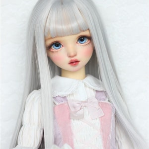 Puede incluir: Una muñeca con el pelo largo, liso, blanco plateado y ojos azules. La muñeca lleva una blusa blanca con cuello de encaje y un vestido rosa y blanco con un lazo.