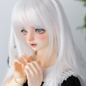 Può includere: Una bambola con i capelli bianchi, gli occhi verdi e il blush rosa sulle guance. Indossa un vestito bianco e nero e tiene le mani giunte davanti a sé.