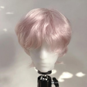 May include: A short, layered wig in a soft pastel pink colour, styled with a voluminous, wavy texture and a side-swept fringe. The wig is displayed on a white mannequin head, showcasing a natural-looking hair fibre with a subtle sheen.