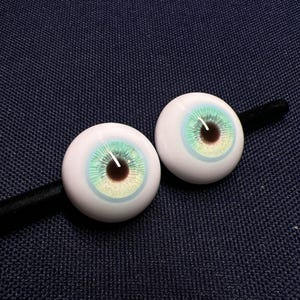 Puede incluir: Dos ojos de mu&ntilde;eca blancos con un iris azul y verde y una pupila marr&oacute;n.