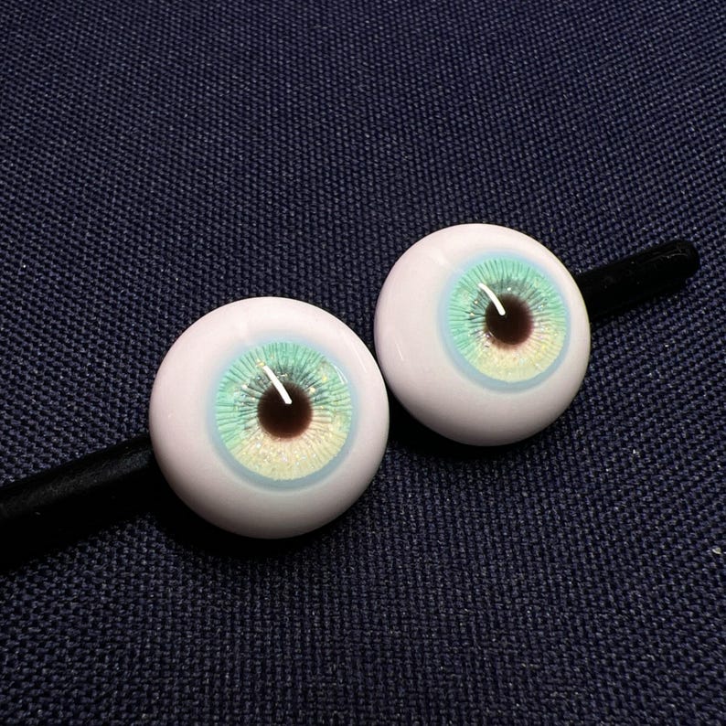 Puede incluir: Dos ojos de mu&ntilde;eca blancos con un iris azul y verde y una pupila marr&oacute;n.
