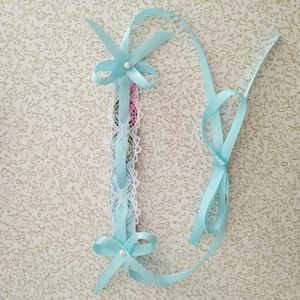 May include: A light blue ribbon with a white lace trim and a pearl accent. The ribbon is tied in a bow at the top and bottom.