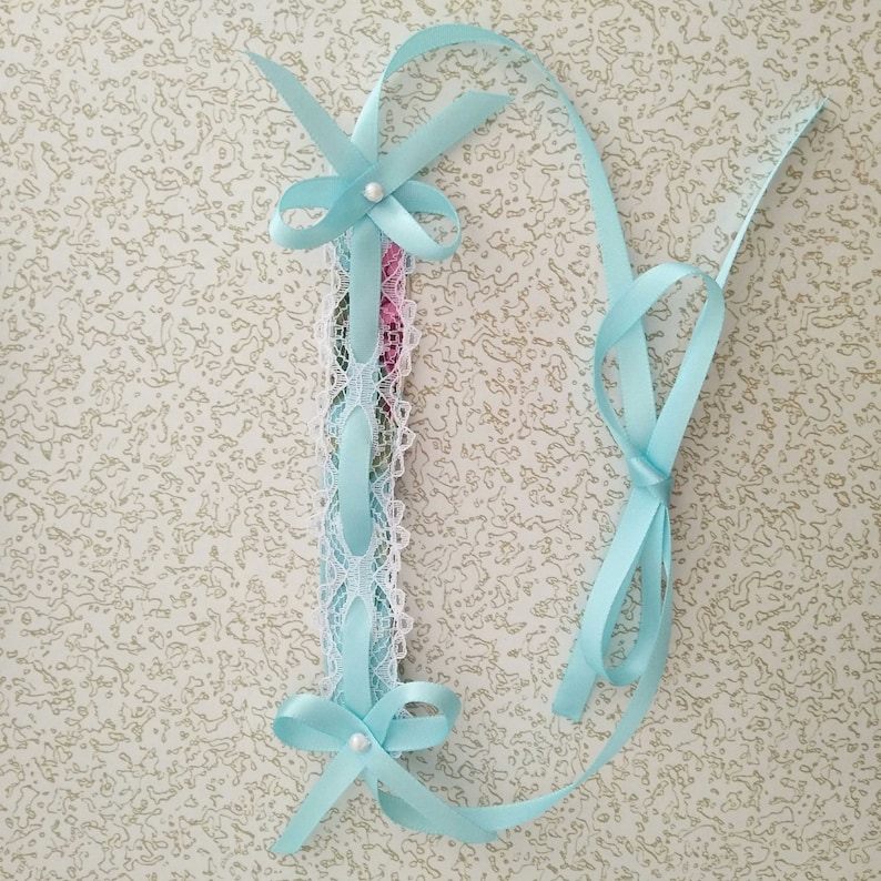 May include: A light blue ribbon with a white lace trim and a pearl accent. The ribbon is tied in a bow at the top and bottom.