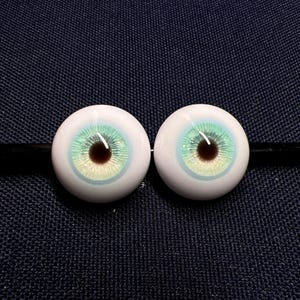 Puede incluir: Dos ojos de mu&ntilde;eca realistas con un iris blanco y una pupila verde y azul. Los ojos est&aacute;n sobre un fondo de tela azul oscuro.