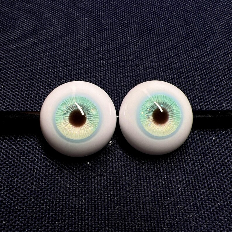 Puede incluir: Dos ojos de mu&ntilde;eca realistas con un iris blanco y una pupila verde y azul. Los ojos est&aacute;n sobre un fondo de tela azul oscuro.