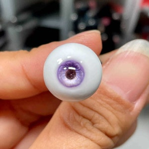 Op de afbeelding: Een close-up van een enkel, wit, poppenoog met een paarse iris en een glanzend, reflecterend oppervlak.