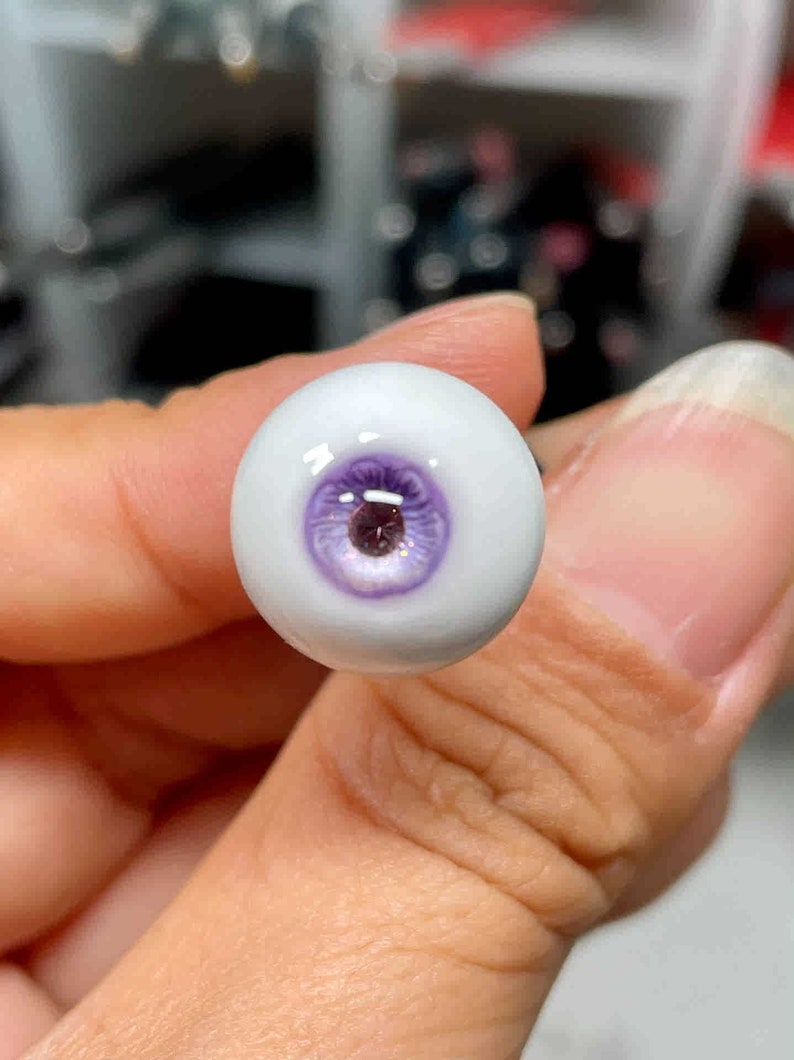 Op de afbeelding: Een close-up van een enkel, wit, poppenoog met een paarse iris en een glanzend, reflecterend oppervlak.