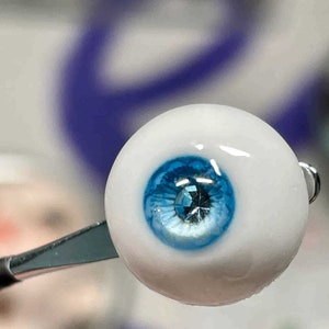 Op de afbeelding: Een close-up van een blauw glazen oog met een heldere, glanzende afwerking. Het oog heeft een klein, helder, glinsterend juweeltje in het midden van de iris.
