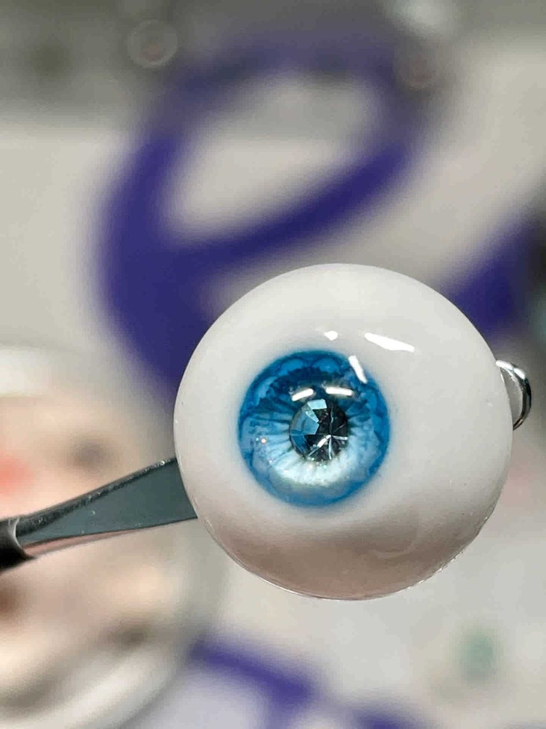 Op de afbeelding: Een close-up van een blauw glazen oog met een heldere, glanzende afwerking. Het oog heeft een klein, helder, glinsterend juweeltje in het midden van de iris.