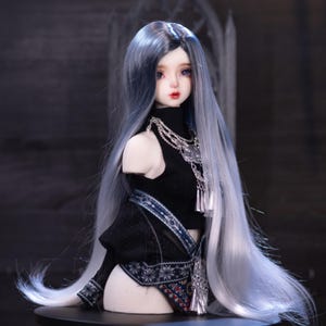 May include: A detailed doll with long, flowing blue-gray hair and delicate facial features. The doll wears a black top with silver necklaces and a decorative skirt with blue and red patterns. The doll is displayed on a black stand.