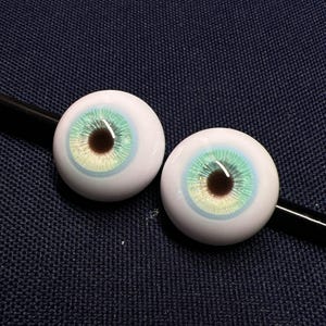 Puede incluir: Dos ojos de vidrio de aspecto realista con una escler&oacute;tica blanca, un iris marr&oacute;n oscuro y una pupila de color azul claro y verde. Los ojos est&aacute;n sobre un fondo de tela azul oscuro.