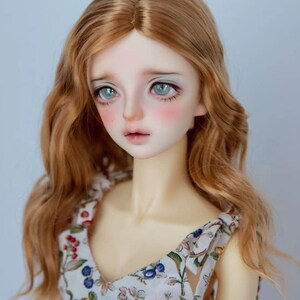 May include: A doll with long, wavy, light brown hair wearing a white dress with a floral pattern. The doll has blue eyes and rosy cheeks.