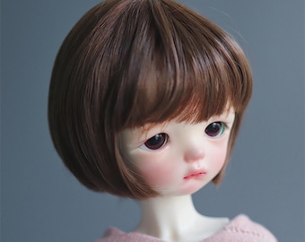 May include: A doll with a pale complexion, rosy cheeks, and dark purple eyes. The doll has short, brown hair styled in a bob cut. It is wearing a pink hooded sweatshirt.
