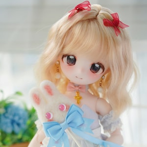 Puede incluir: Una muñeca con cabello rubio y ojos azules que lleva un vestido azul claro y un juguete de peluche de conejo blanco. La muñeca lleva lazos rojos en el pelo y pendientes de oro.