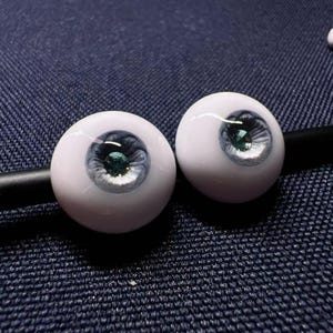 Op de afbeelding: Twee witte poppenogen met een blauwe iris en een groene edelsteenpupil.