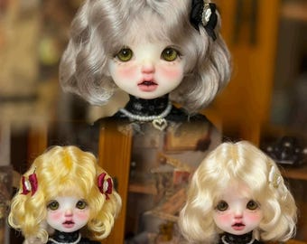 May include: Three dolls with different colored wigs. The dolls have light skin, green eyes, and are wearing black dresses. The wigs are blonde, light blonde, and silver.