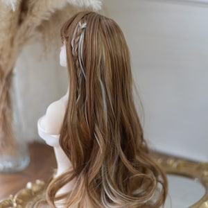 May include: A long, wavy wig with a mix of brown and blonde highlights. The wig features a side braid with white strands woven in. The hair is styled on a mannequin head, with a soft, blurred background.