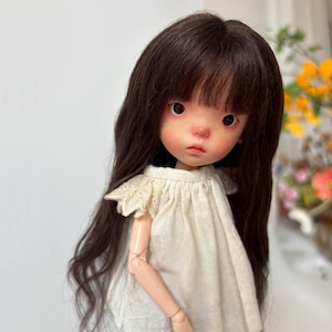 May include: A doll with long, dark brown hair and a fair complexion. The doll has large eyes, a small nose, and a slightly downturned mouth. It wears a cream-colored dress with lace detailing on the shoulders.
