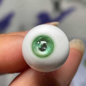 Op de afbeelding: Een close-up van een groen glazen oog met een witte sclera. Het oog wordt vastgehouden door een vinger.