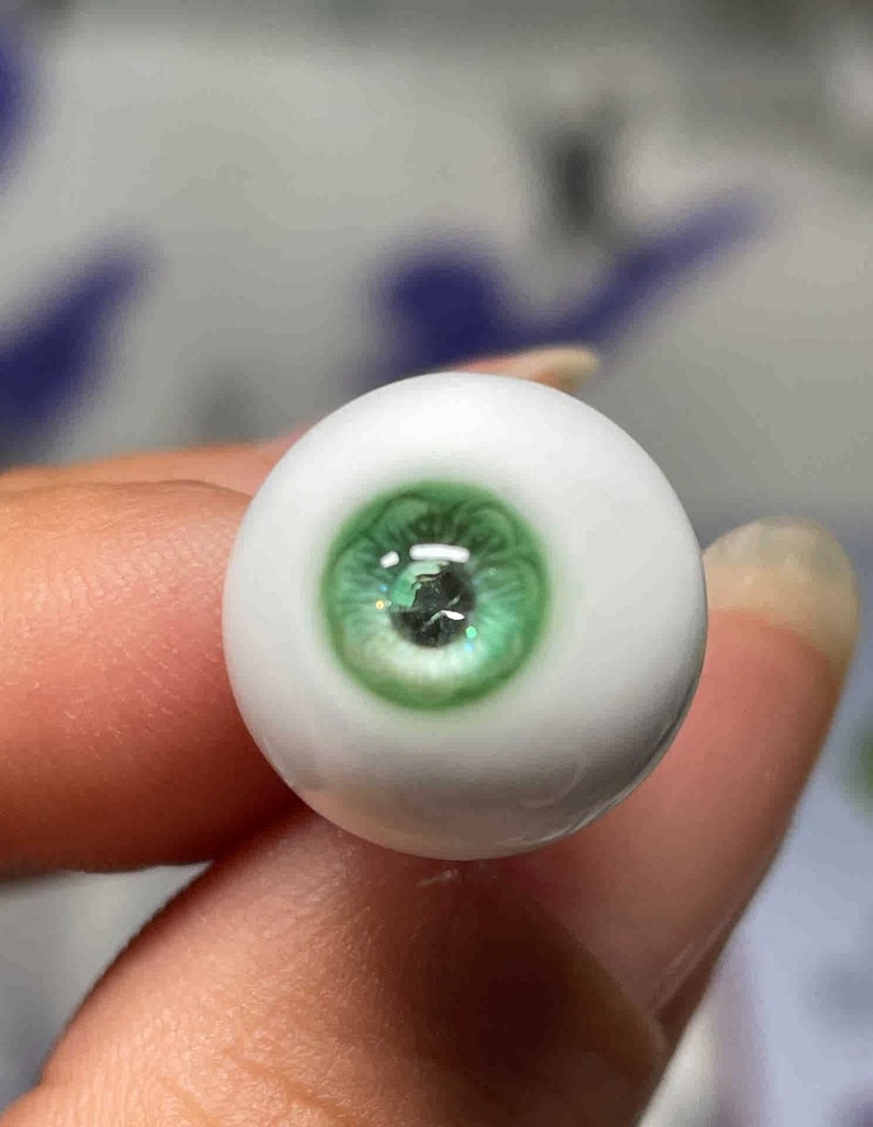 Op de afbeelding: Een close-up van een groen glazen oog met een witte sclera. Het oog wordt vastgehouden door een vinger.