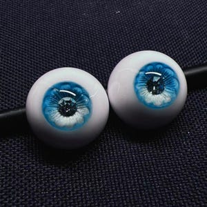 Op de afbeelding: Twee blauwe en witte poppenogen met een glinsterend centrum. De ogen zijn rond en hebben een witte rand rond de blauwe iris.