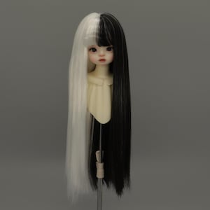 May include: A doll head with long, straight hair, split evenly into black and white sections. The doll has a pale complexion, delicate facial features, and dark eyes. The doll is mounted on a stand against a neutral gray background.