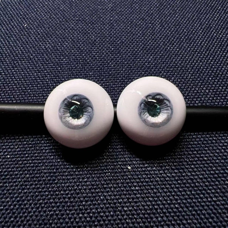 Op de afbeelding: Twee witte poppenogen met een blauwe en groene iris en een zwarte pupil.