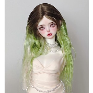 Può includere: Una bambola con capelli lunghi e ondulati, castani alle radici e che sfumano al verde alle punte. La bambola indossa un vestito bianco e una collana di perle.