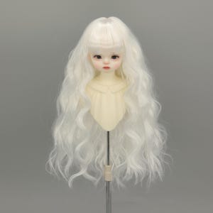 May include: A doll head with a pale complexion, delicate facial features, and long, wavy white hair. The doll's face has subtle makeup, including defined eyebrows and rosy lips. The head is attached to a light-colored torso.