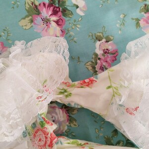 May include: White lace trim with ruffled edges against a blue background with a floral pattern. The floral pattern features pink, white, and green roses.