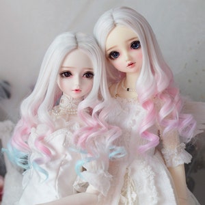 May include: Two dolls with long, white wigs with pink and blue ombre highlights. The wigs are styled in loose curls. The dolls are wearing white dresses with lace details.