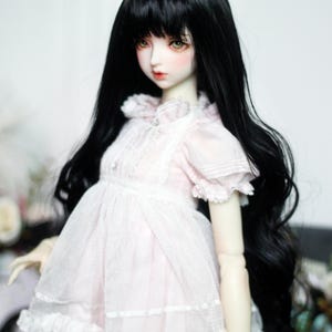 May include: A doll with long, black hair and a pale complexion. The doll is wearing a light pink dress with white lace detailing. The dress has short sleeves and a ruffled skirt. The doll's eyes are a light brown color.