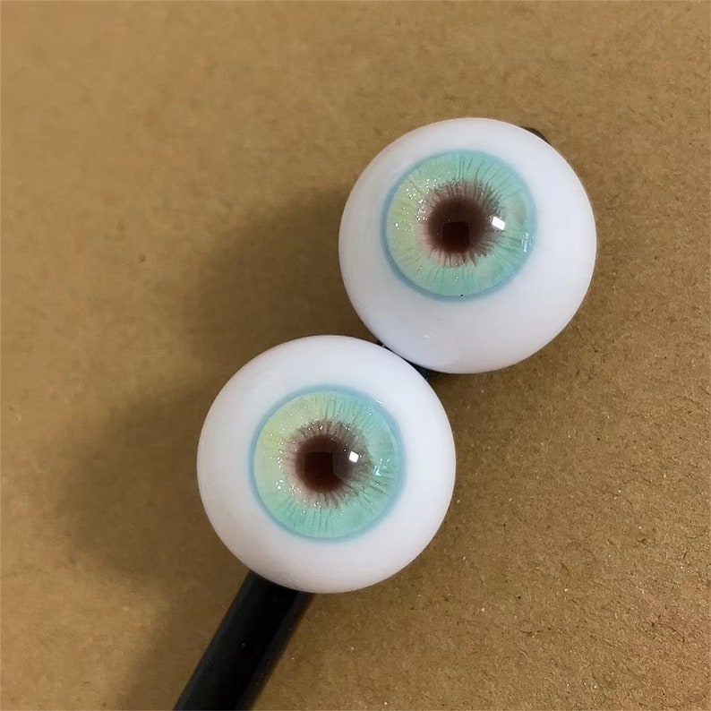 Puede incluir: Dos ojos de mu&ntilde;eca blancos con un iris azul y verde y una pupila marr&oacute;n.