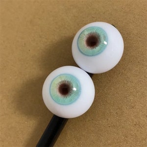 Puede incluir: Dos ojos redondos, blancos, de mu&ntilde;eca con iris azul y verde y pupila marr&oacute;n. Los ojos est&aacute;n en un palo negro.