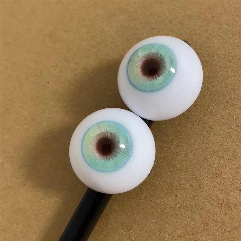 Puede incluir: Dos ojos redondos, blancos, de mu&ntilde;eca con iris azul y verde y pupila marr&oacute;n. Los ojos est&aacute;n en un palo negro.