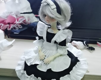 May include: A doll dressed in a black and white maid outfit with a ruffled skirt and sleeves. The doll has gray hair and a black headband. The doll is sitting on a wooden surface. Other doll clothes are visible in the background.