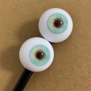 Puede incluir: Dos ojos de mu&ntilde;eca blancos con iris azules, verdes y marrones. Los ojos est&aacute;n en un palo negro.
